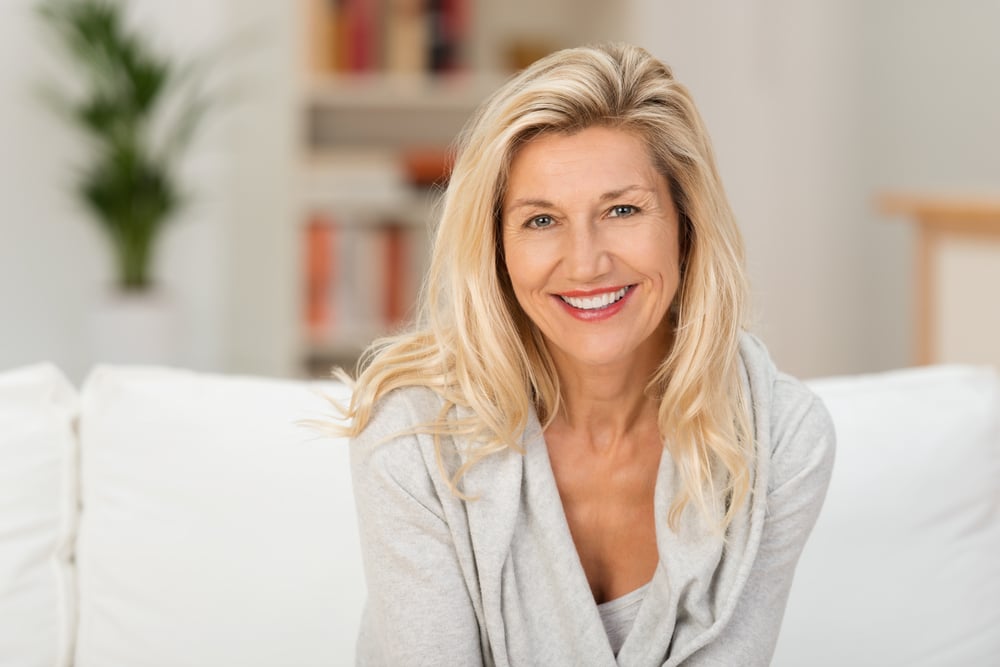 Lovely middle-aged blond woman with a beaming smile sitting on a sofa at home looking at the camera-1